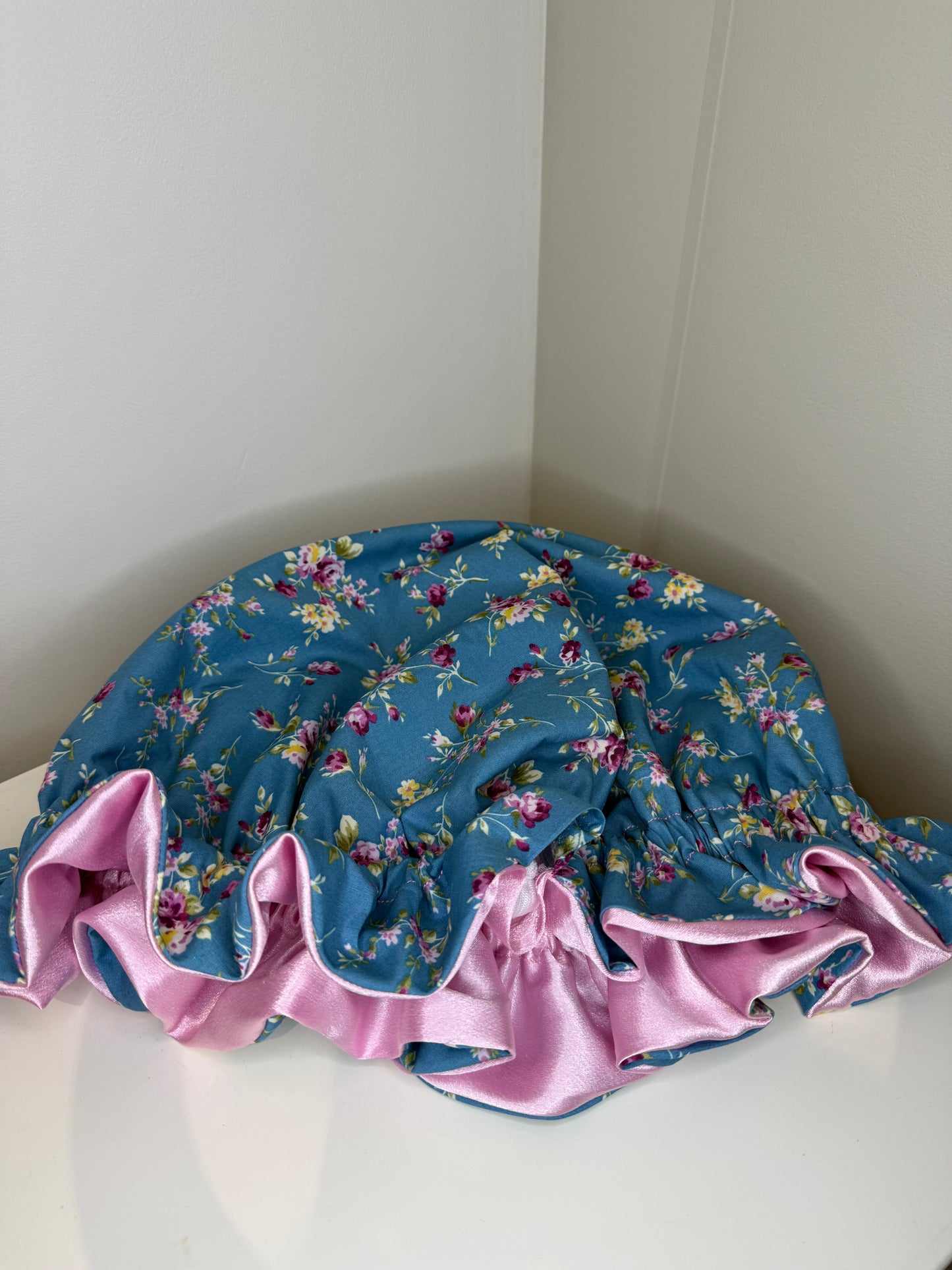 A flat satin ruffle bonnet with a blue floral print and a pink satin ruffled trim, resting on a white surface in a corner. It shows the adjustable toggle inside, the shiny pink interior and the beautiful design on the exterior.
