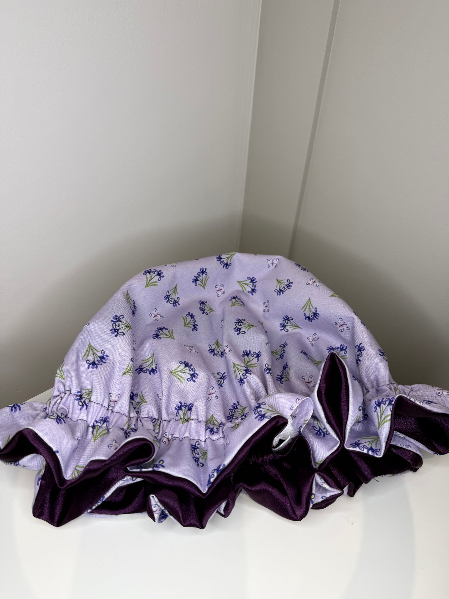 A lavender satin sleep cap with a purple floral print and a dark purple ruffled trim, displayed on a white surface in a corner.