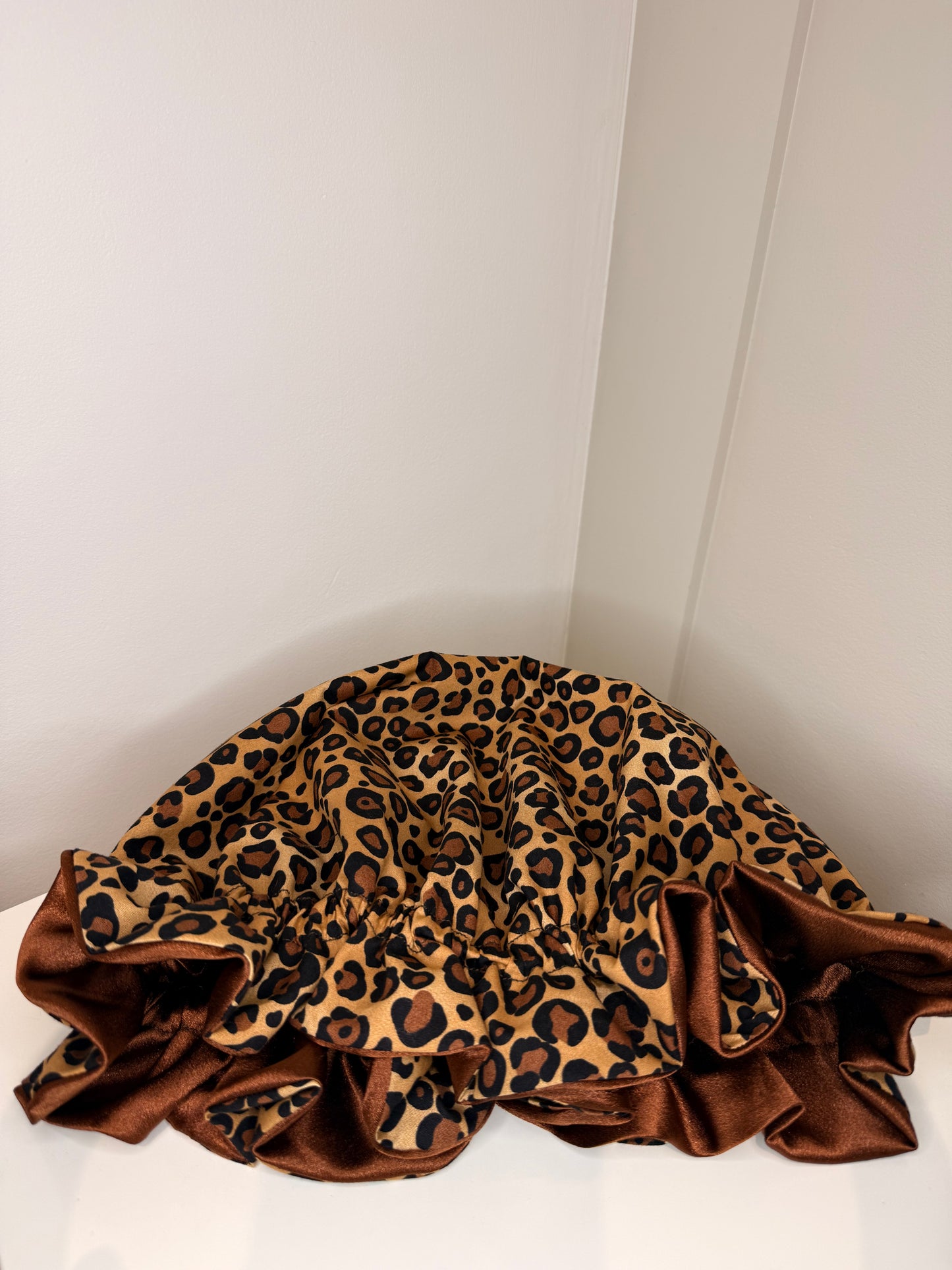 A leopard print satin bonnet with a brown ruffled border, draped on a white surface against a white wall corner.