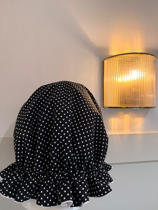 Black polka dot bonnet on a mannequin against a neutral wall with a wall-mounted lamp.