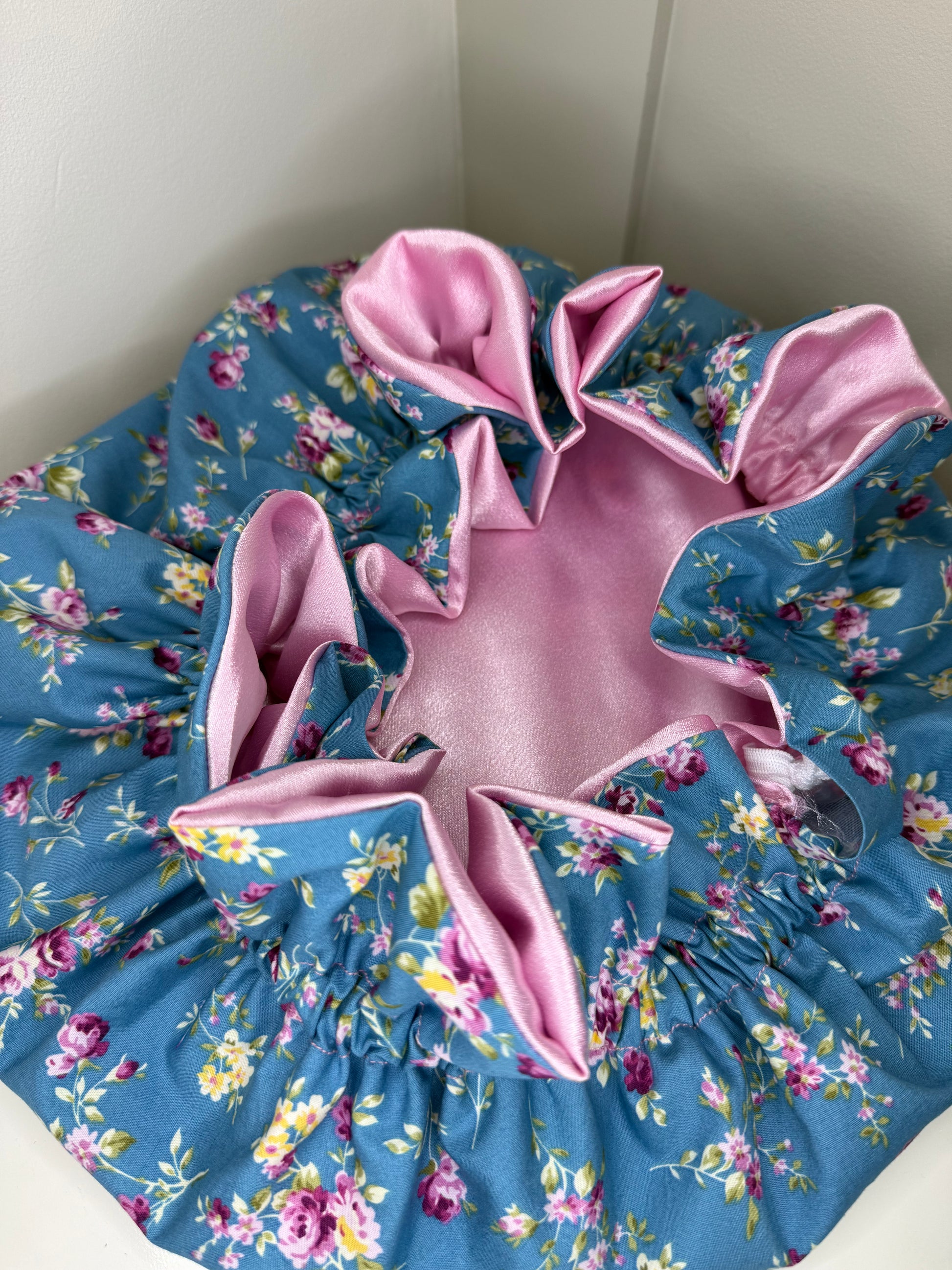 A cute bonnet with a blue floral print exterior and a shiny pink satin interior, photographed in a home setting against a light-colored wall.