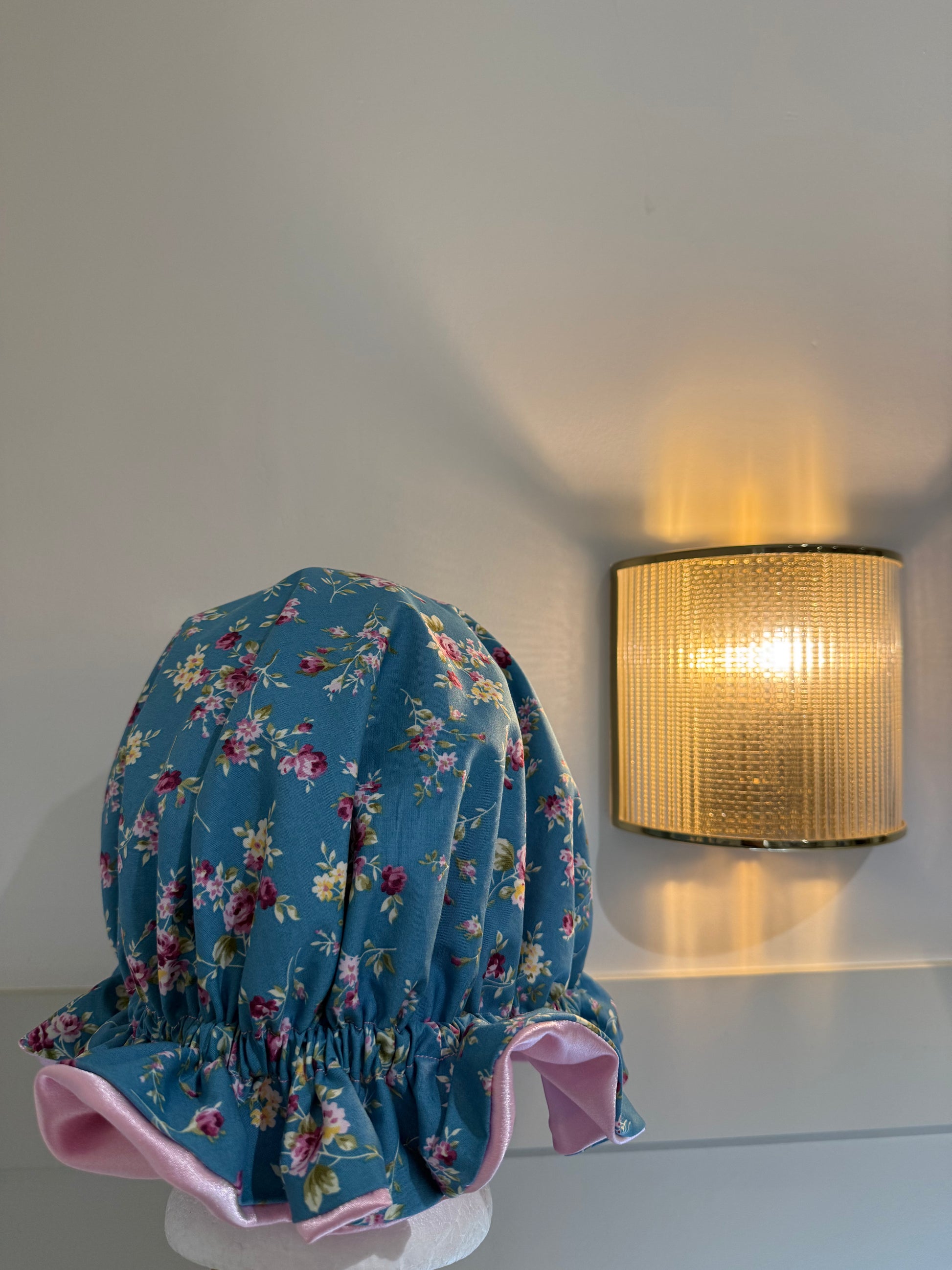 A blue floral bonnet with pink ruffled trim displayed on a mannequin head against a light gray wall, with a lit wall sconce visible in the background.