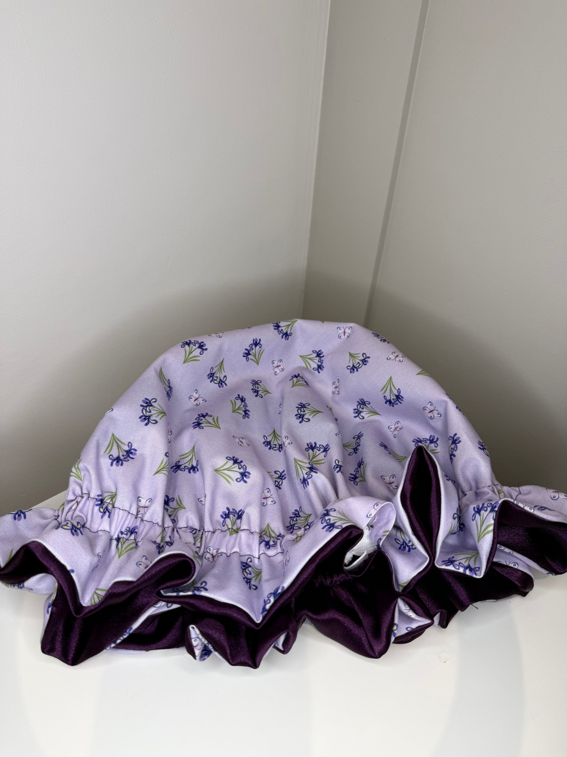 A lavender satin sleep cap with a purple floral print and a dark purple ruffled trim, displayed on a white surface in a corner.