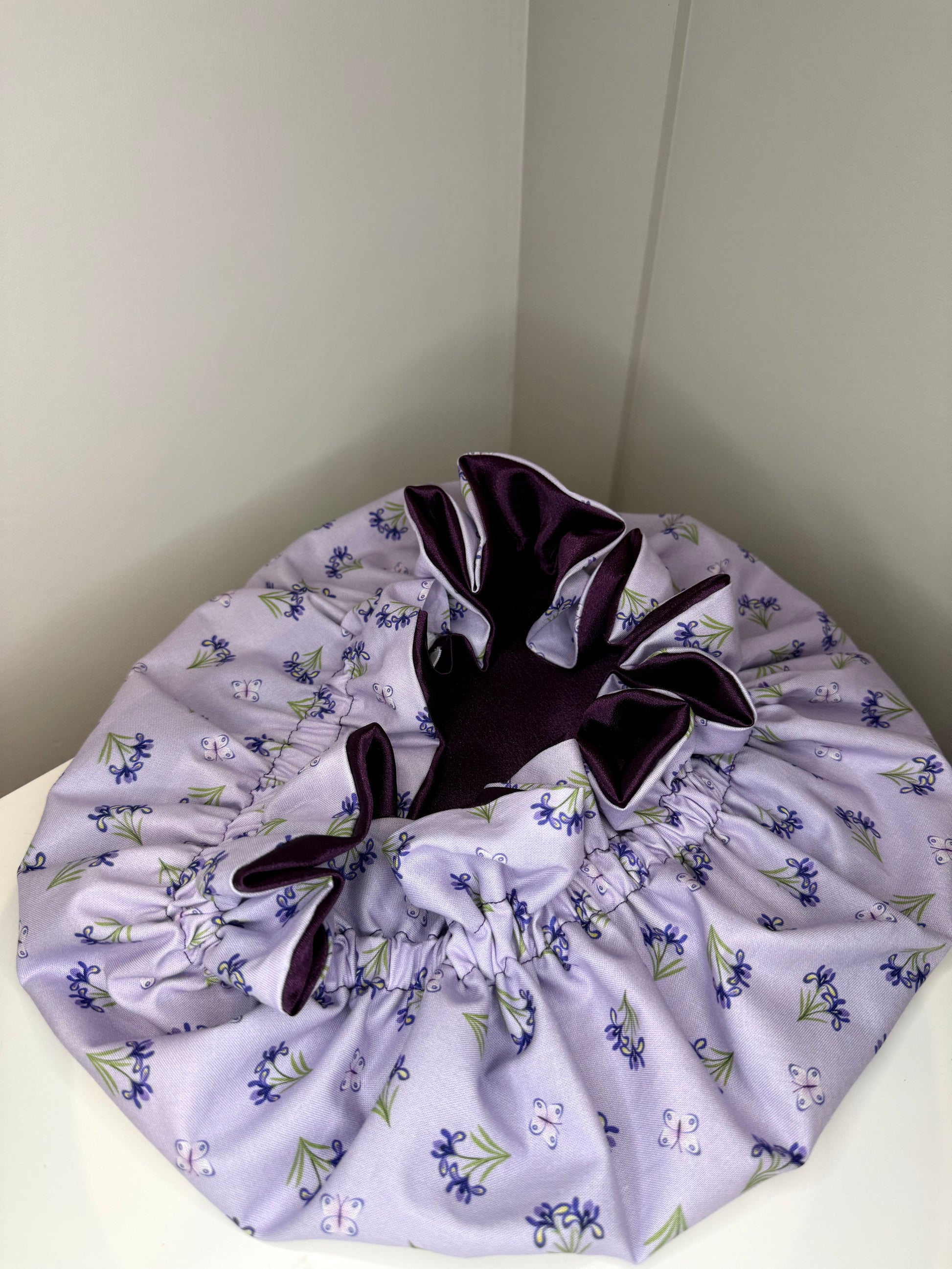 A lavender drawstring bonnet with a floral print of purple flowers and green leaves, featuring a dark purple satin lining and drawstring closure.