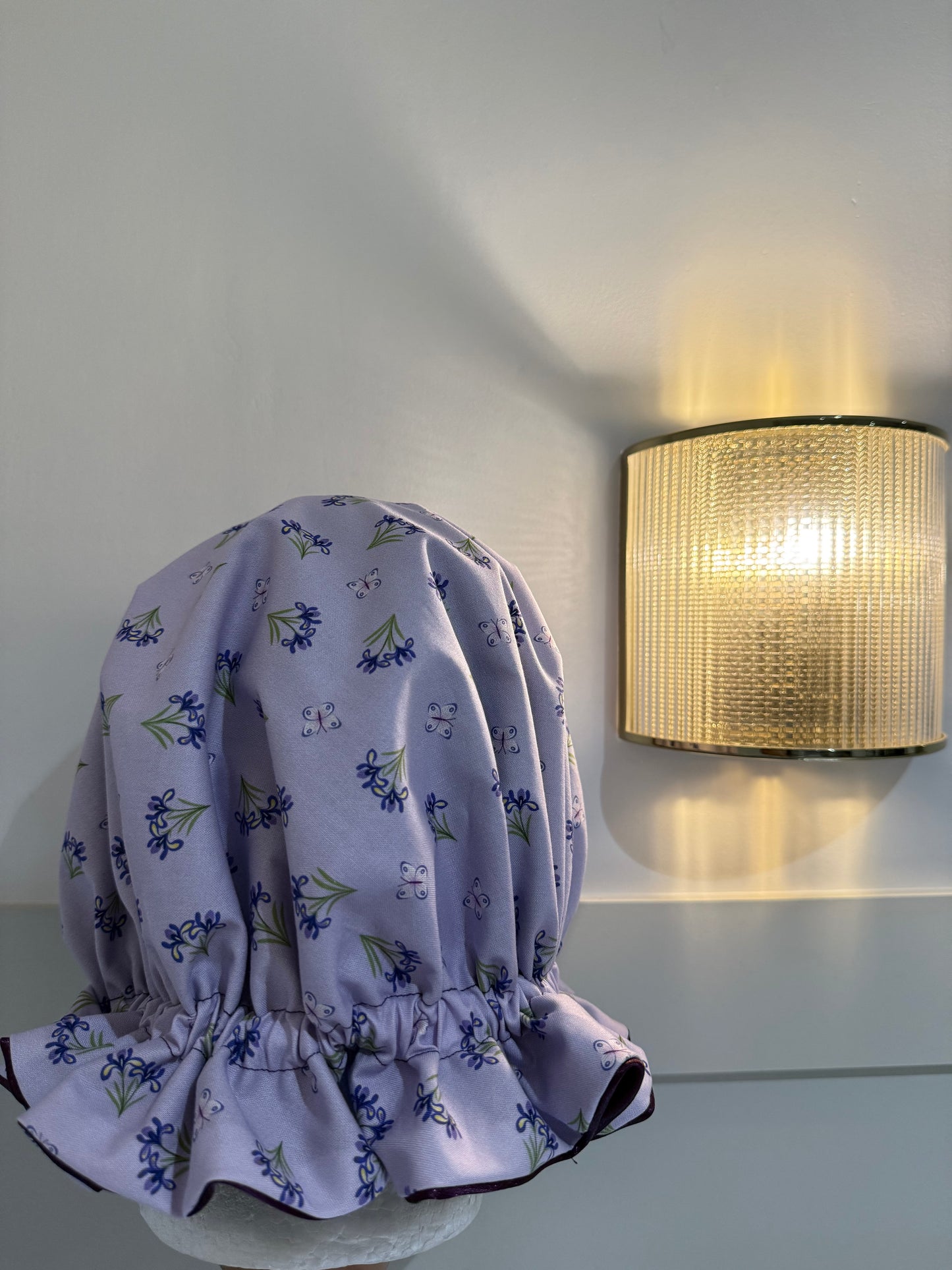 A lavender bonnet with a purple and green floral print, displayed on a mannequin head against a light gray wall next to a glowing cylindrical wall lamp.