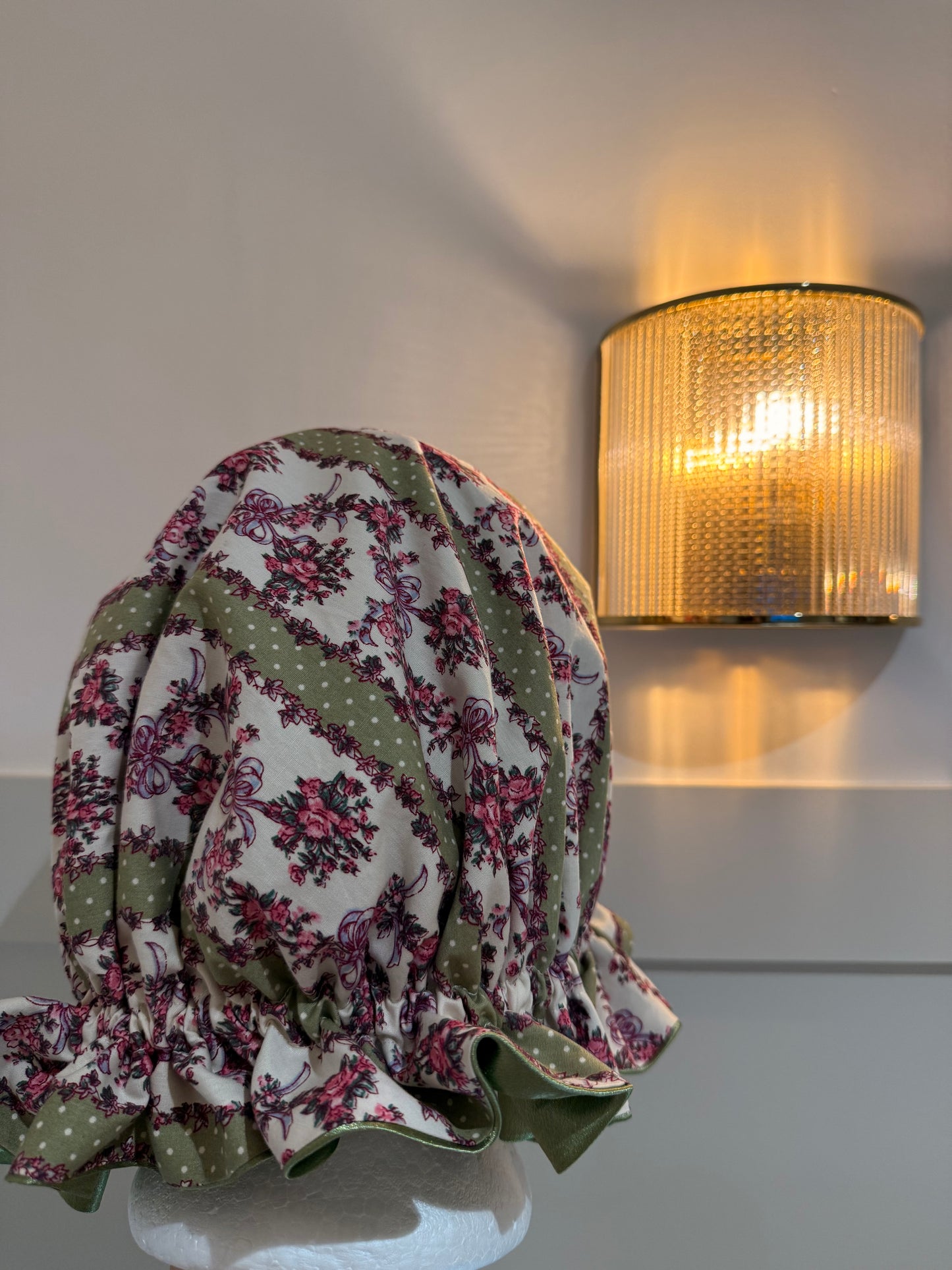  floral bonnet with bows and polka dots, with sage green satin lining on a white surface next to a wall-mounted light fixture.