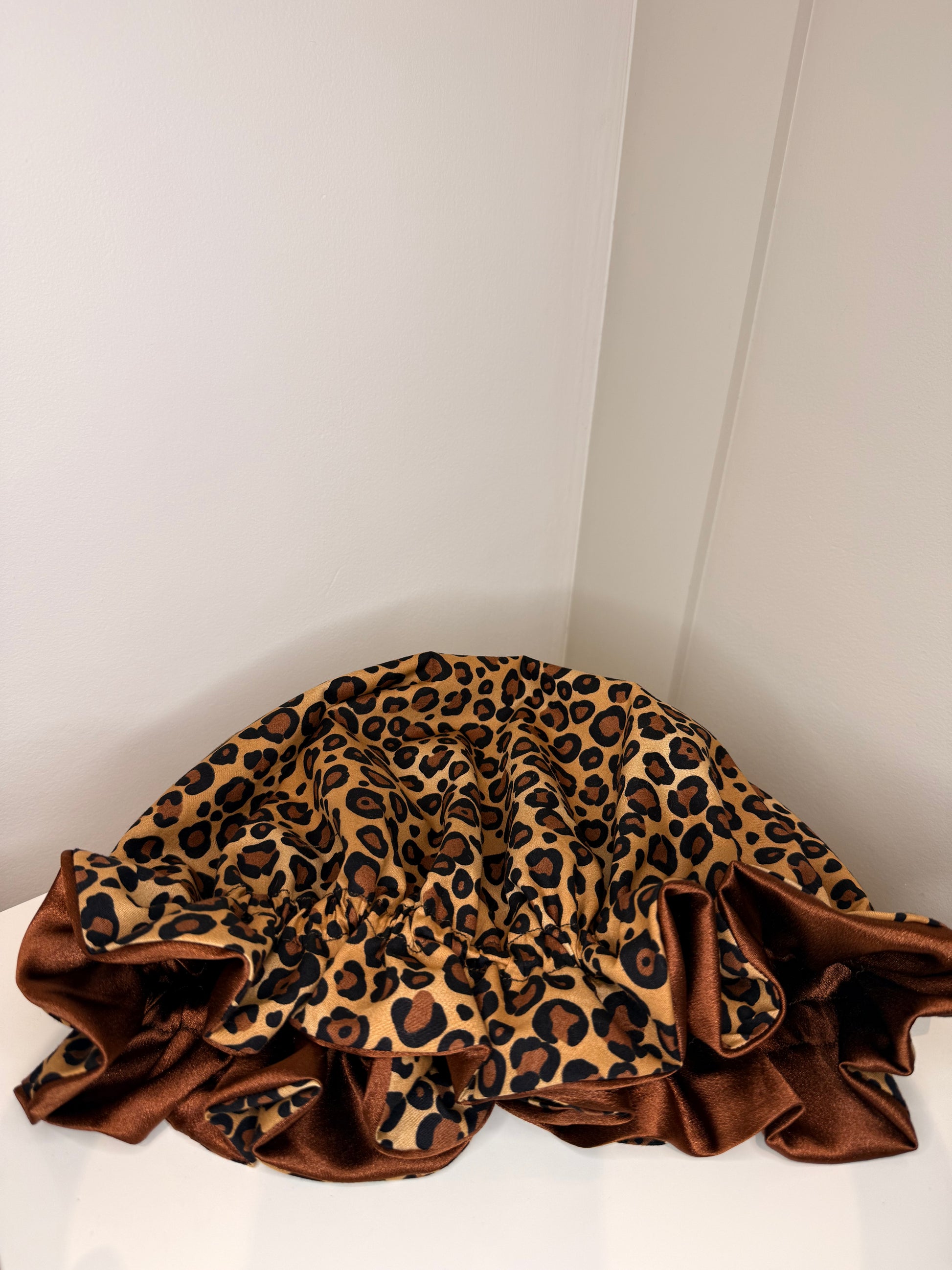 A leopard print satin bonnet with a brown ruffled border, draped on a white surface against a white wall corner.