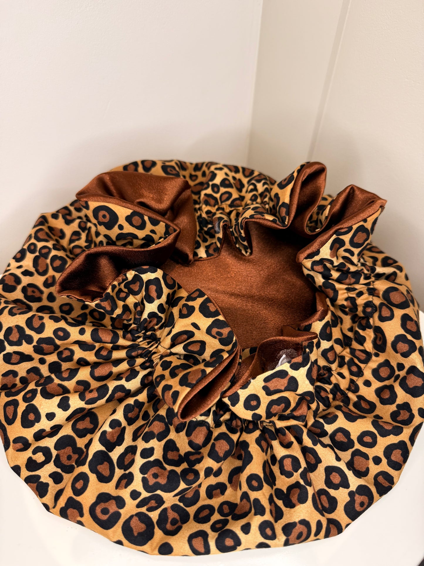 A leopard print satin adjustable bonnet with a brown satin inner lining, photographed in the corner of a white room.