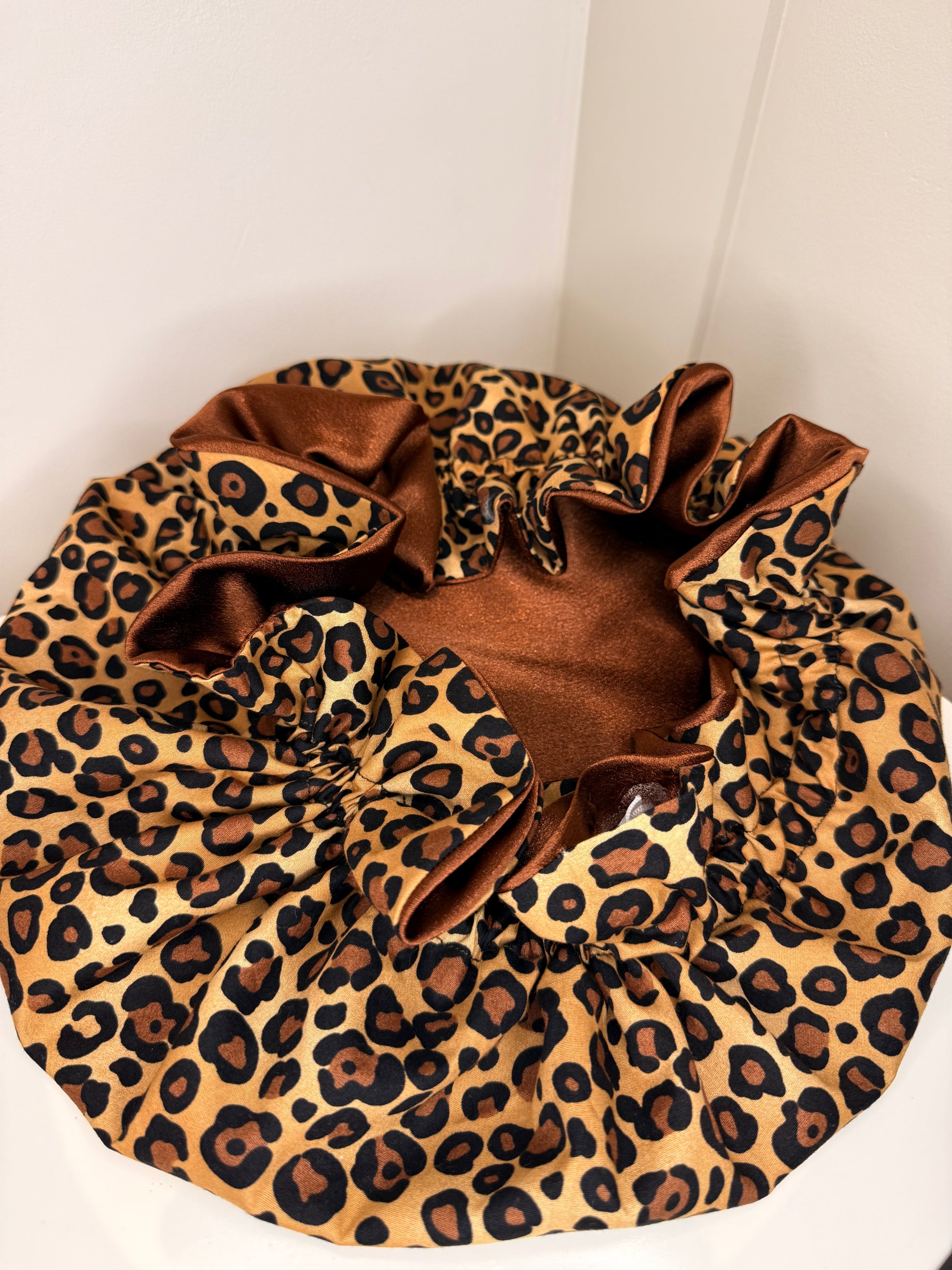 A leopard print satin adjustable bonnet with a brown satin inner lining, photographed in the corner of a white room.