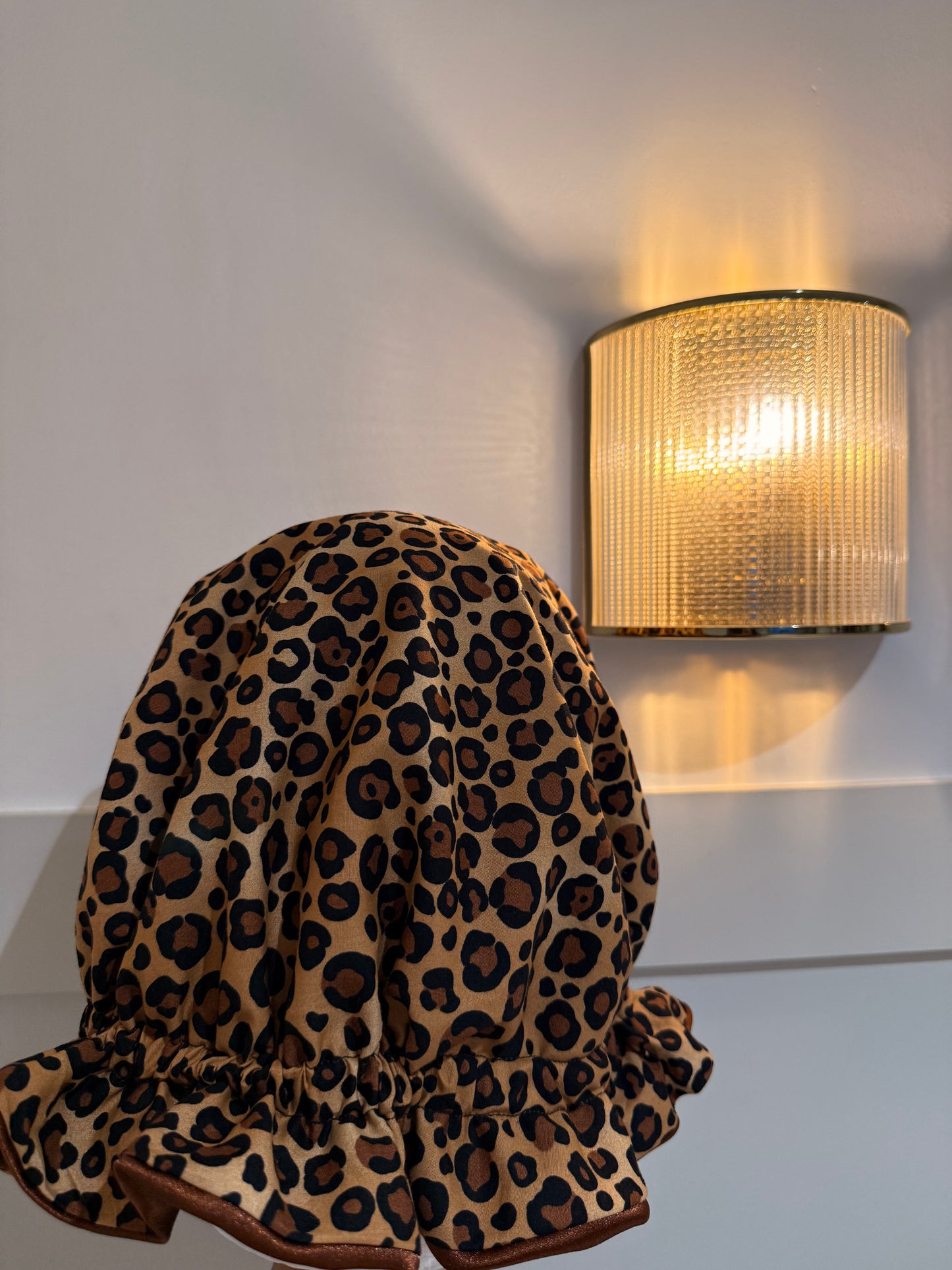 A leopard print fabric satin bonnet draped over a mannequin head in a home setting, with a warm glowing wall source on the white wall behind it.