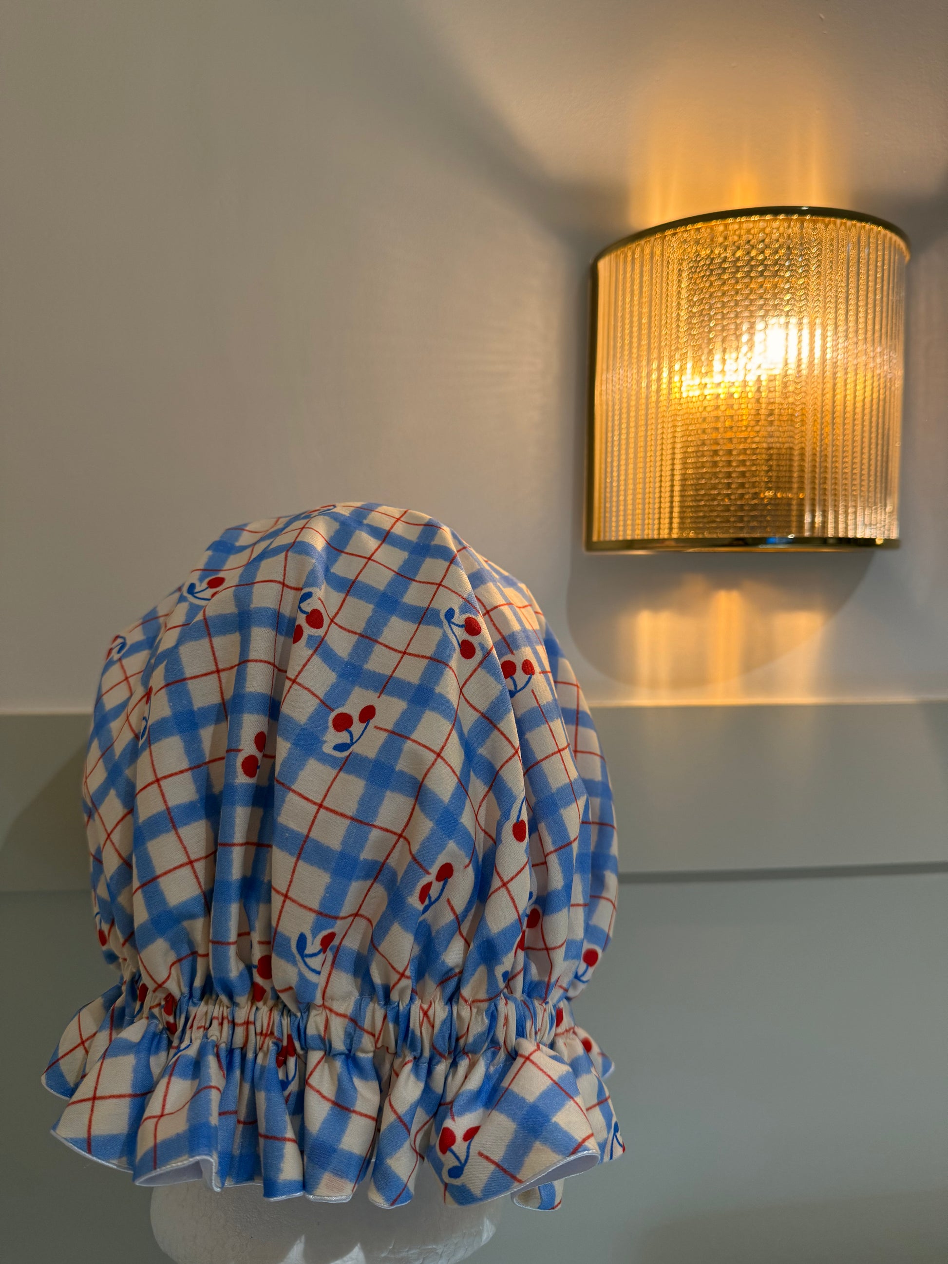 Blue and white checkered satin bonnet with red cherries on a mannequin against a wall with a gold light fixture.