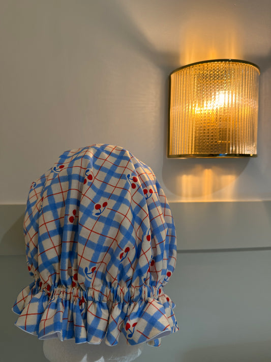 Blue and white checkered satin bonnet with red cherries on a mannequin against a wall with a gold light fixture.
